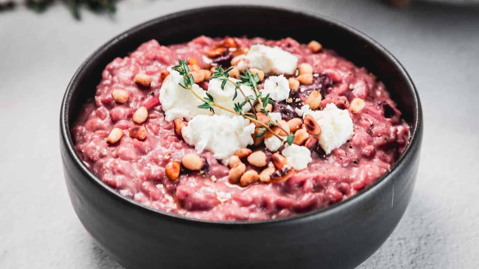 A black bowl filled with beetroot risotto, topped with pine nuts, goat cheese, and a small sprig of thyme, placed on a light-colored surface.