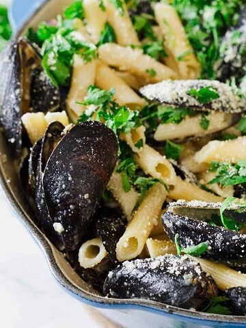 A close-up of a blue pot filled with cooked penne pasta, mussels in shells, chopped parsley, and grated cheese, with some fresh parsley scattered around the pot on a white surface.