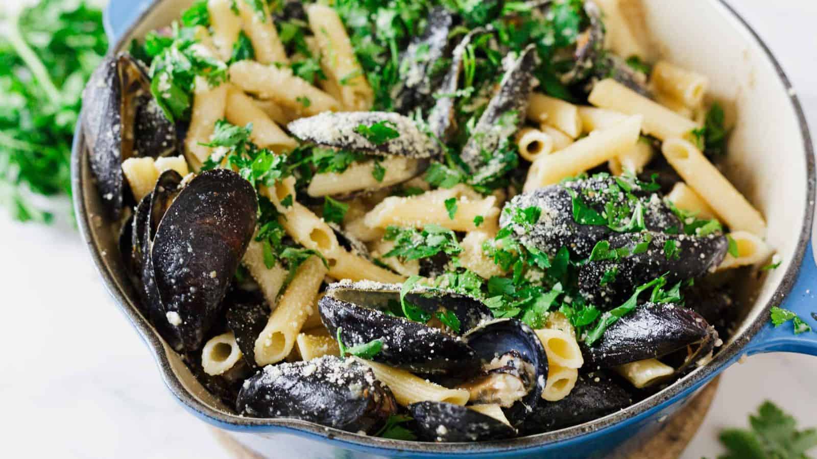 A close-up of a blue pot filled with cooked penne pasta, mussels in shells, chopped parsley, and grated cheese, with some fresh parsley scattered around the pot on a white surface.