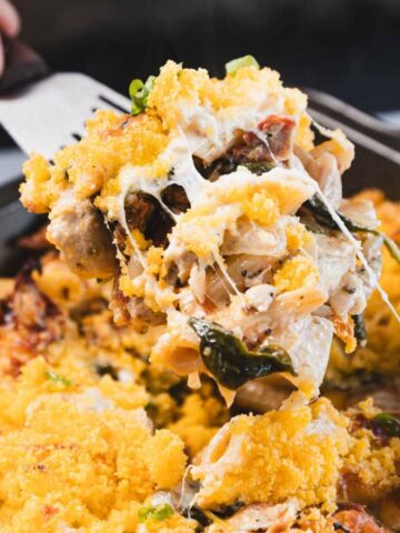 A close-up of a hand lifting a cheesy pasta bake with vegetables from a baking dish using a spatula. The dish has a golden, crumbly topping and visible melted cheese.