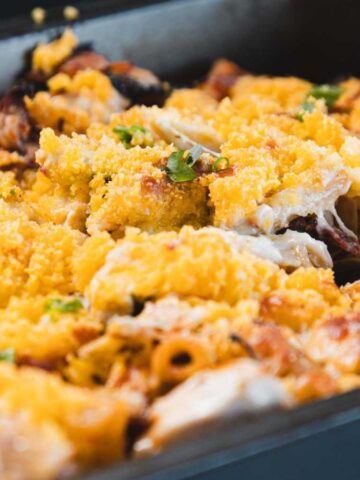 A close-up of a baked pasta dish in a rectangular metal pan with a golden breadcrumb topping, pieces of pasta, baked cheese, and green herbs visible.