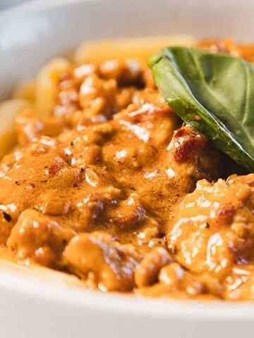 A close-up of a bowl of rigatoni pasta topped with a creamy meat sauce and garnished with two fresh basil leaves.