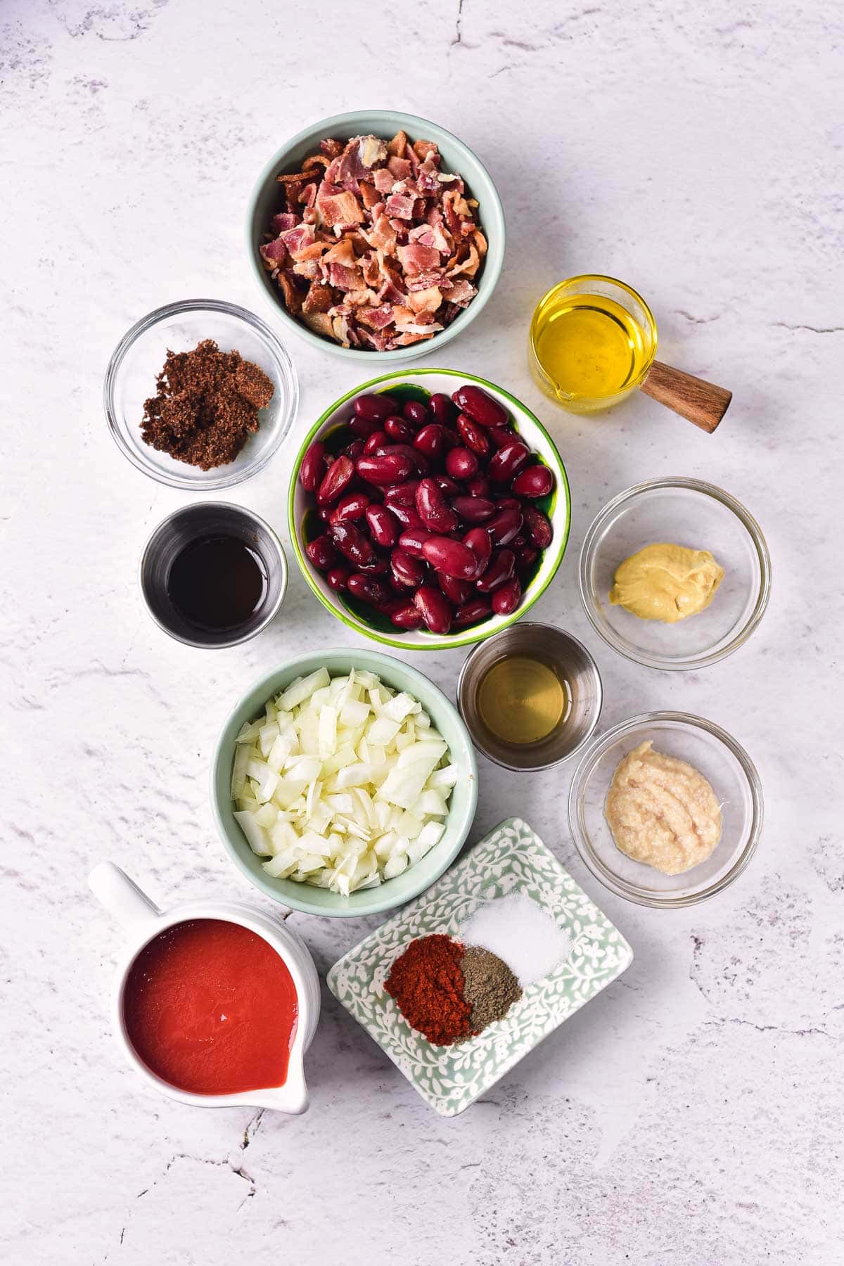 An overhead view of small bowls with ingredients for Slow Cooker Baked Beans With Bacon&mdash;kidney beans, chopped bacon, onion, brown sugar, tomato sauce, spices, and more&mdash;arranged on a white surface for this delicious slow cooker recipe.