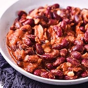 A white bowl filled with slow cooker baked beans in a tomato-based sauce sits on a dark textured cloth, with green herbs blurred in the background.