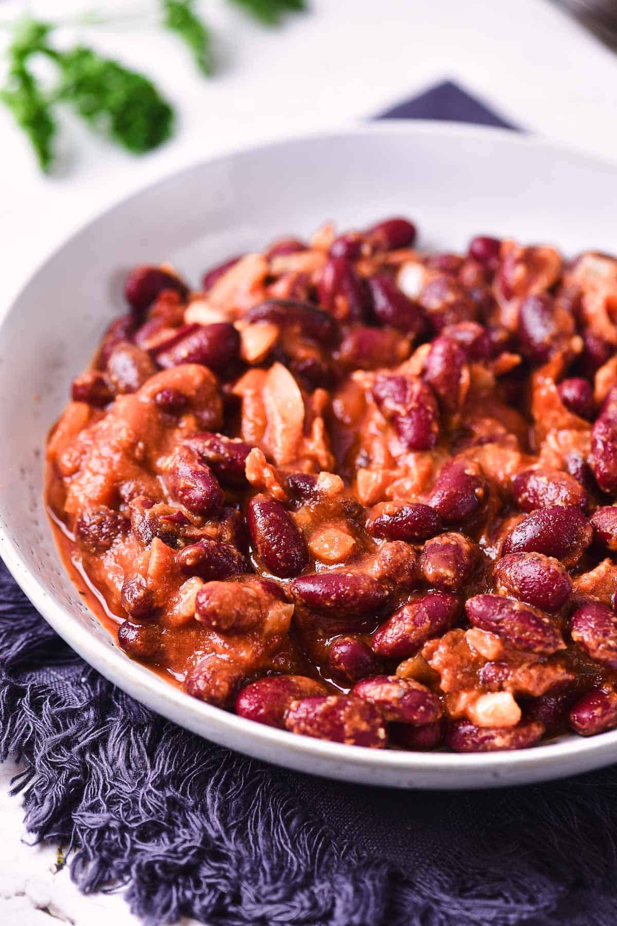 A white bowl filled with slow cooker baked beans in a tomato-based sauce sits on a dark textured cloth, with green herbs blurred in the background.