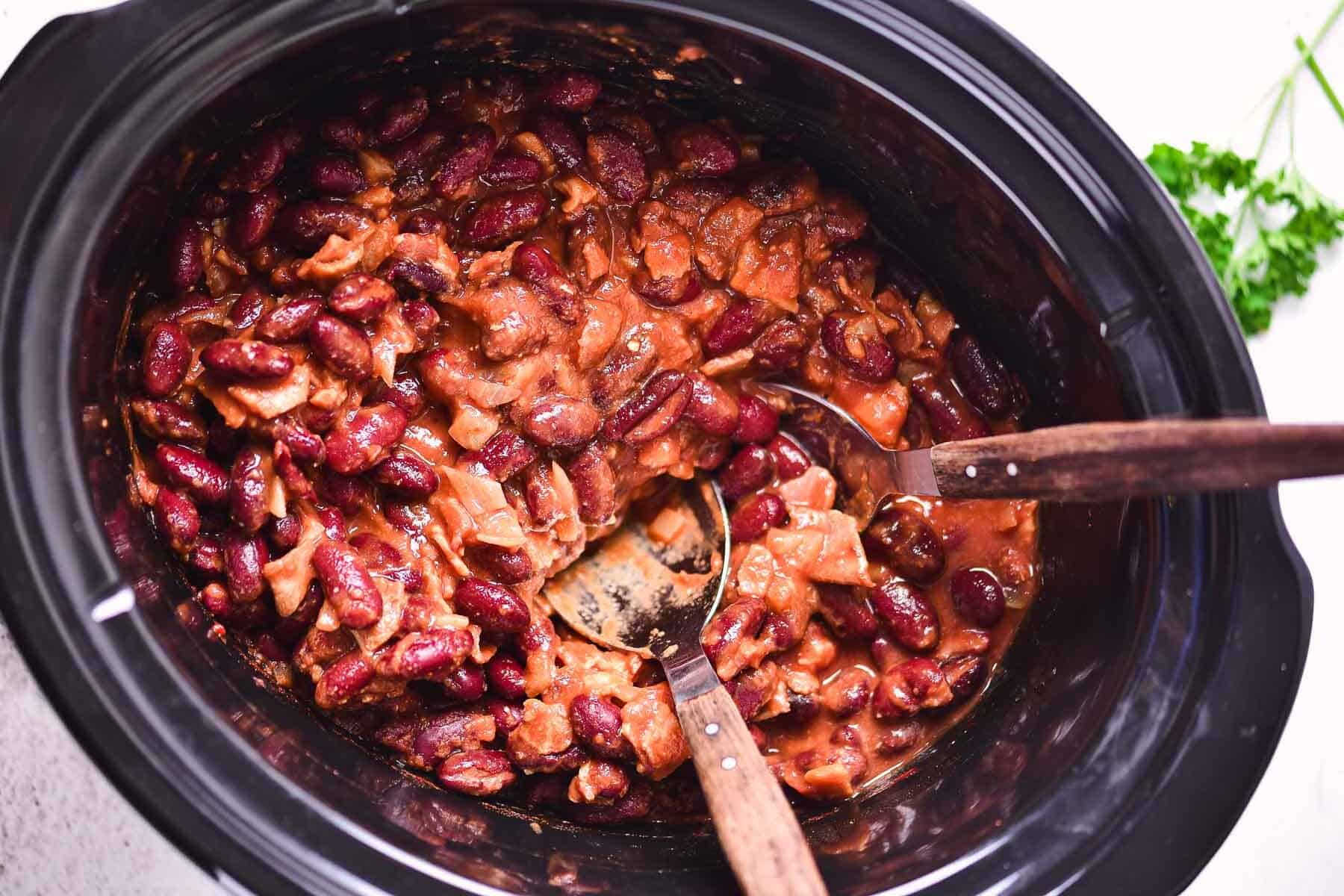 A slow cooker filled with chili containing red kidney beans, chunks of meat, and sauce brings cozy comfort. Two wooden-handled serving spoons rest inside, while a sprig of parsley sits on the white countertop&mdash;perfect inspiration for a hearty bacon recipe.