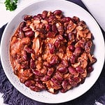 A white bowl filled with Slow Cooker Baked Beans in a thick reddish-brown sauce, with visible pieces of onion and bacon, placed on a dark cloth napkin.