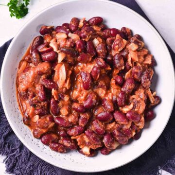 A white bowl filled with Slow Cooker Baked Beans in a thick reddish-brown sauce, with visible pieces of onion and bacon, placed on a dark cloth napkin.