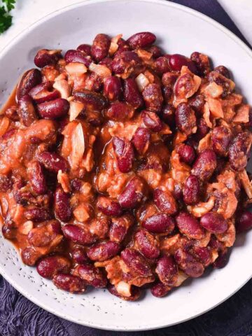 A white bowl filled with Slow Cooker Baked Beans in a thick reddish-brown sauce, with visible pieces of onion and bacon, placed on a dark cloth napkin.