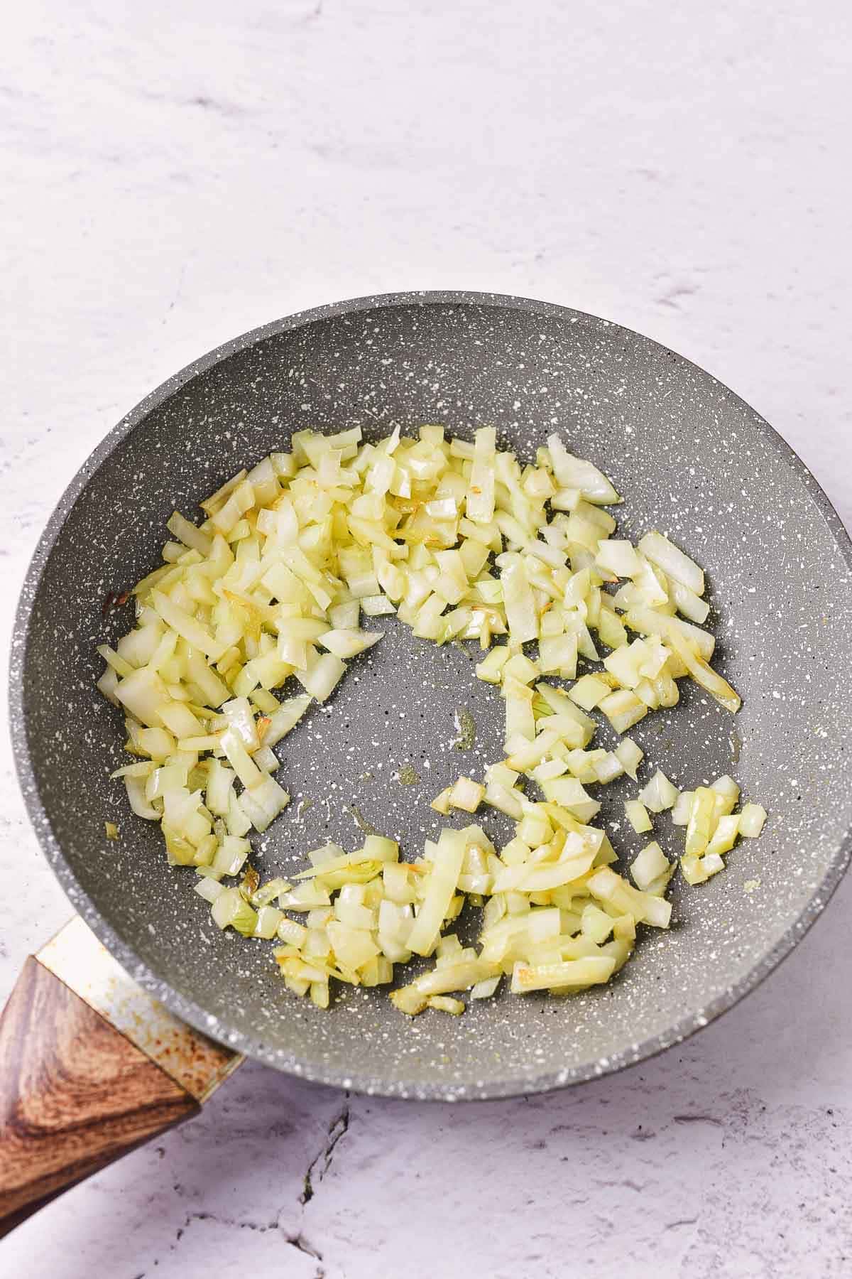 Chopped onions are being saut&eacute;ed in a nonstick frying pan with a wooden handle on a light-colored surface, perfect as a base for a hearty baked beans recipe.