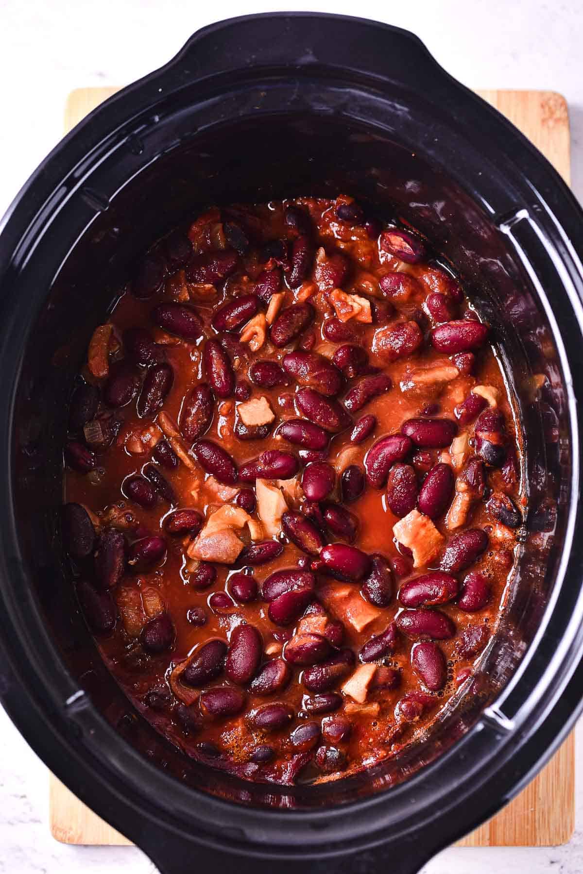 A slow cooker filled with a hearty stew of red kidney beans and savory meat in a rich reddish-brown sauce, perfect for a comforting Slow Cooker Baked Beans dish, set on a wooden surface.