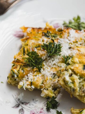 A slice of vegetable frittata topped with grated cheese and parsley sits on a floral plate. A fork is positioned beside the frittata, and another piece of parsley lies on the plate.
