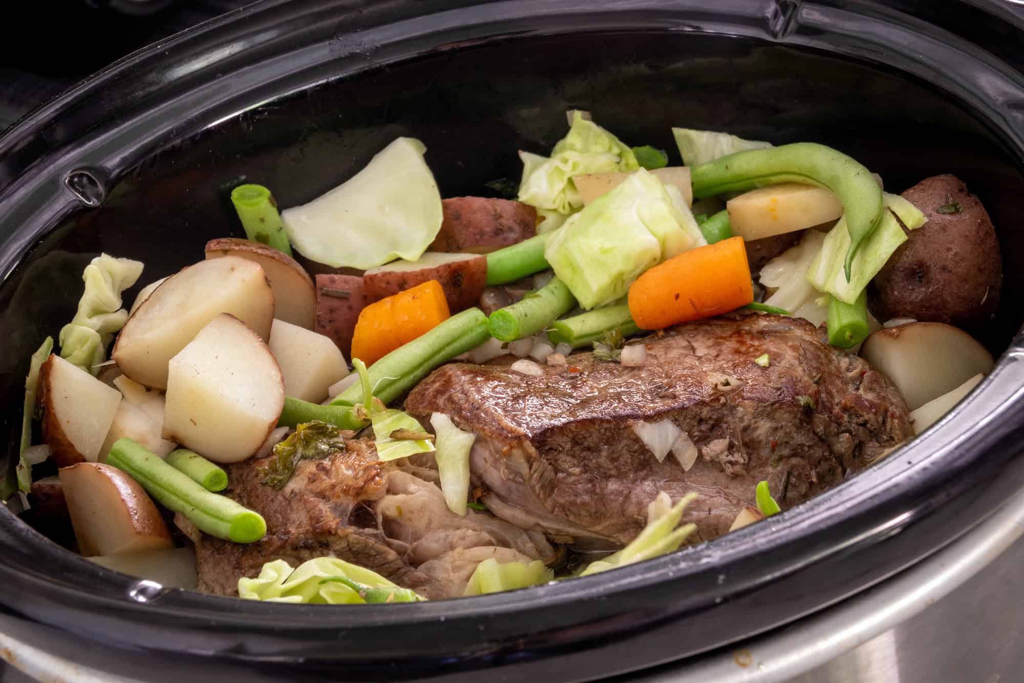 A slow cooker filled with a pot roast, tender potatoes, carrots, green beans, and cabbage&mdash;juices pooling at the bottom. This is everything you need to know for perfect results every time.