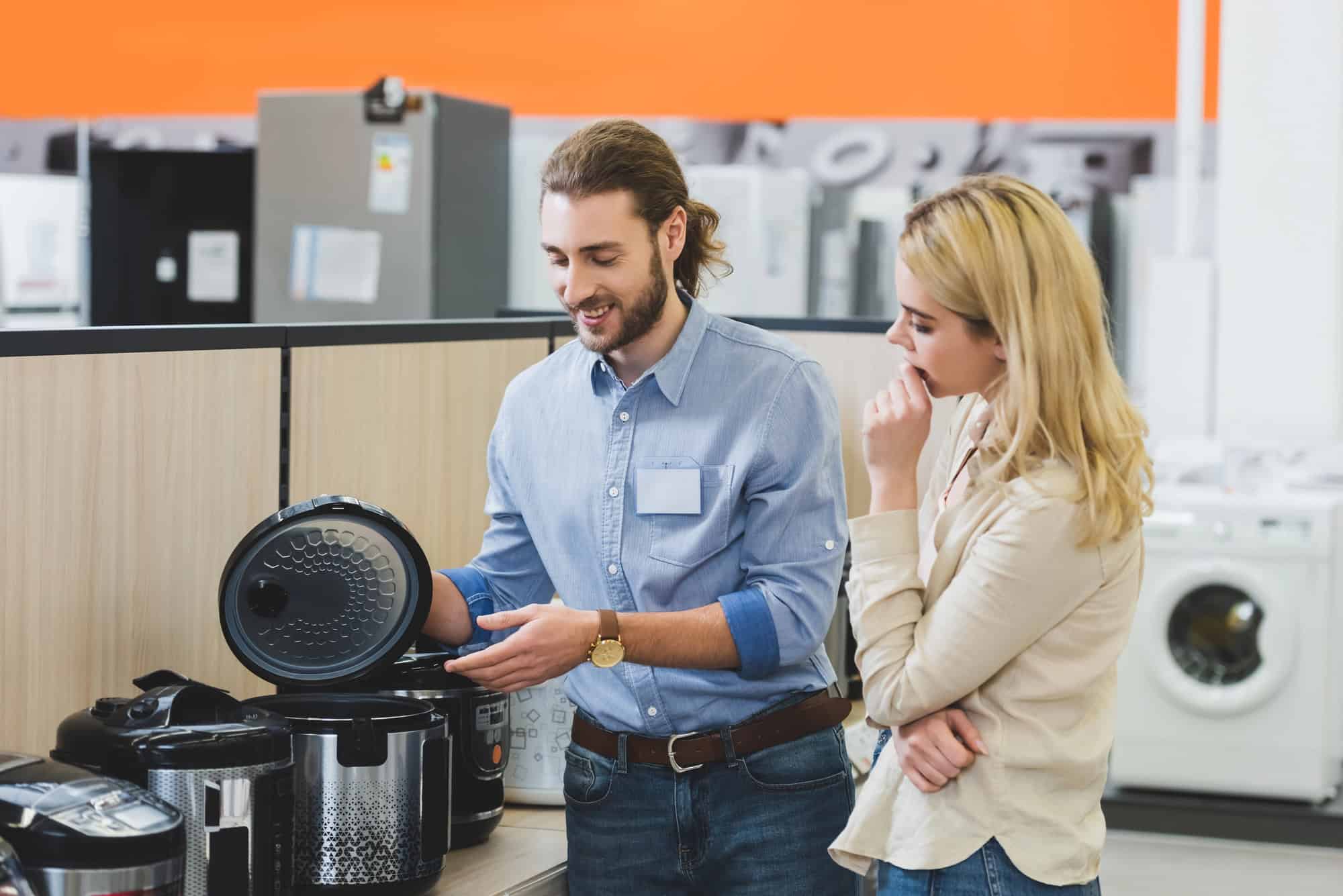 A man in a blue shirt shows a rice cooker with the lid open to a woman in a beige blouse at an appliance store, sharing slow cooker tips as washing machines and refrigerators are visible in the background.