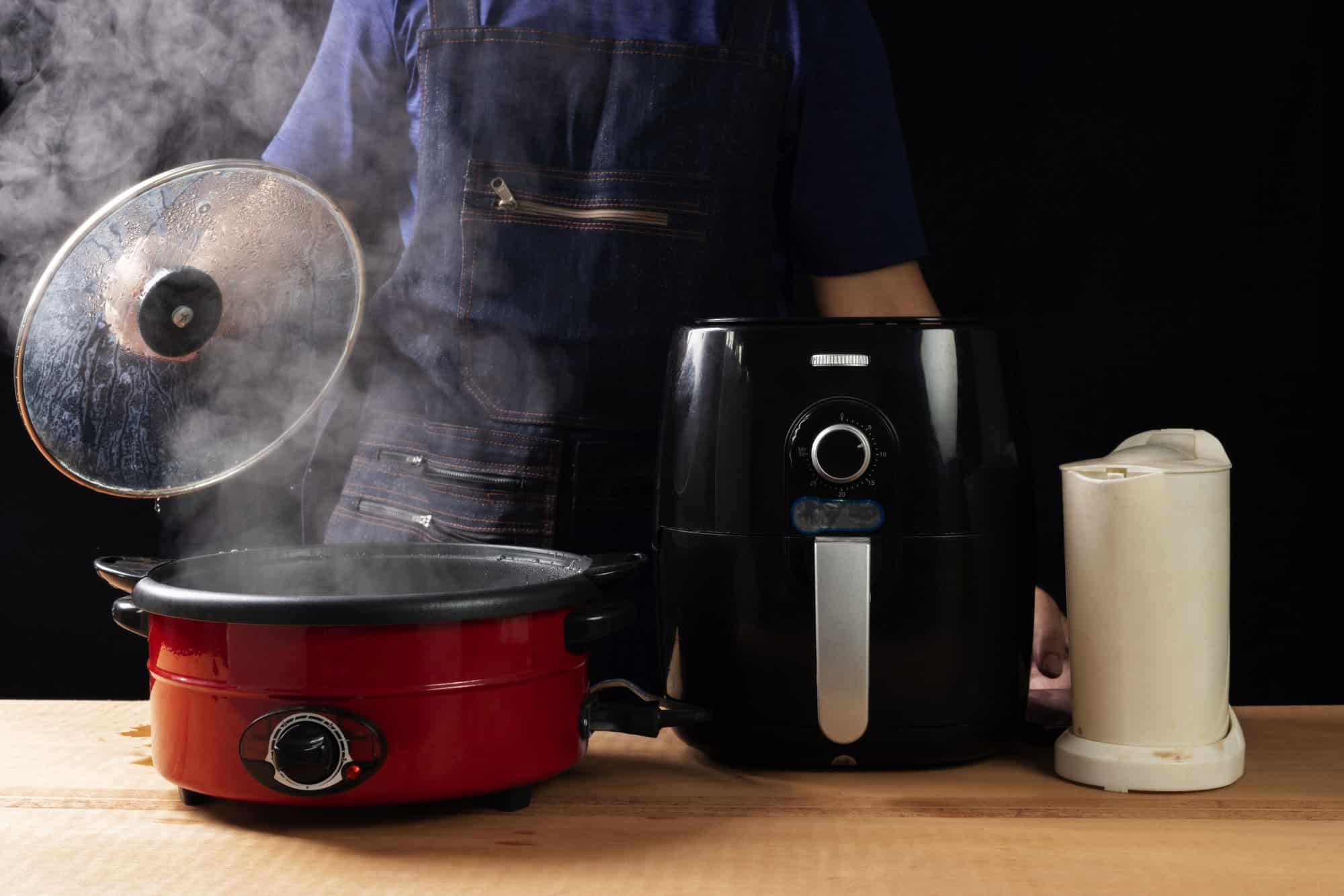 A person wearing a blue apron lifts the lid off a steaming red slow cooker. An air fryer and an electric kettle sit on a wooden table next to the slow cooker, creating the perfect scene for sharing handy slow cooker tips.