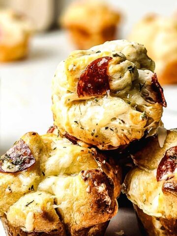 Three muffin-shaped pieces of pull-apart bread with melted cheese and pepperoni are stacked on a plate. A small bowl of dip and more bread pieces are visible in the background.