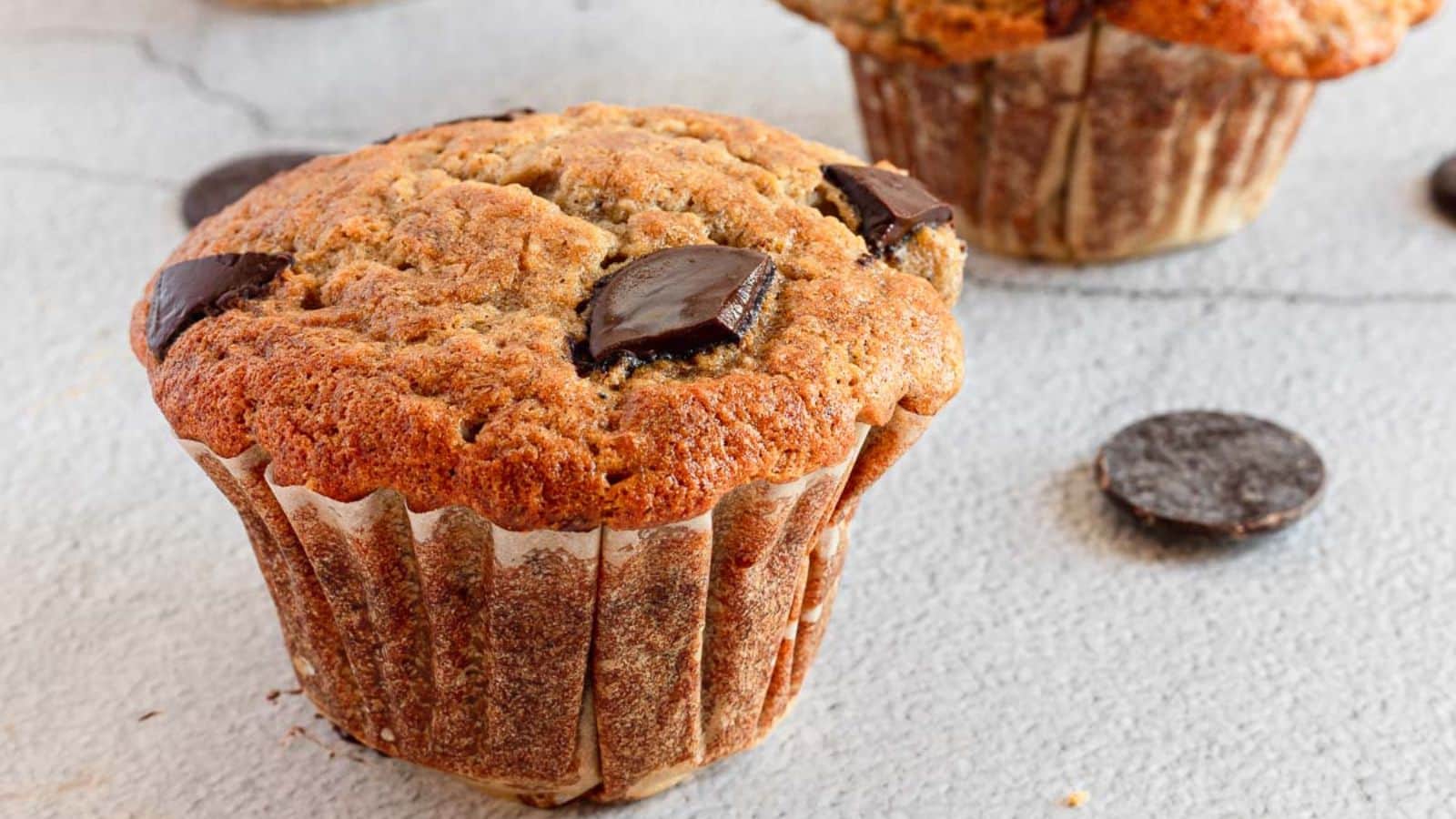 A close-up of a muffin with visible chocolate chunks, wrapped in a brown paper liner, sits on a light textured surface with another muffin and some chocolate pieces in the background.