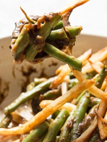 A casserole dish filled with cooked green beans and thin, crispy French fries, with a spoon lifting a portion of the mixture. Some fries and beans are coated in a dark sauce.