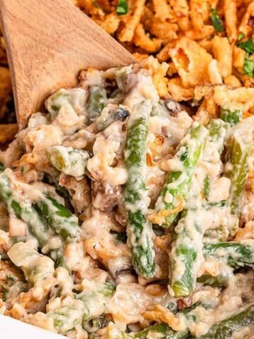 A close-up of a green bean casserole in a white baking dish, topped with crispy fried onions. A wooden spoon is lifting a portion, showing green beans mixed with creamy sauce and mushrooms.
