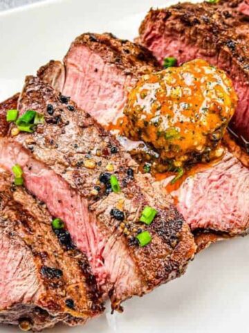 Three thick slices of medium-rare steak are arranged on a white plate, garnished with chopped chives and topped with a dollop of seasoned butter.