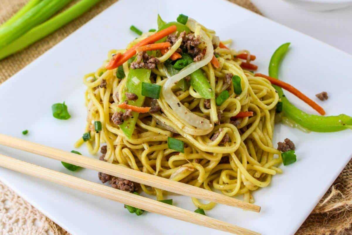 A plate of stir-fried noodles with vegetables and beef, garnished with green onions, and a pair of chopsticks on the side.