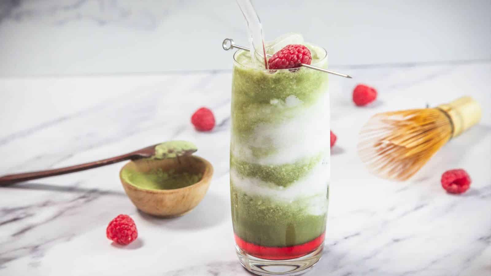 A tall glass with swirled layers of green and white iced drink, topped with two raspberries on a metal pick. Surrounding the glass are loose raspberries, a wooden spoon with green powder, a small bowl, and a bamboo whisk.