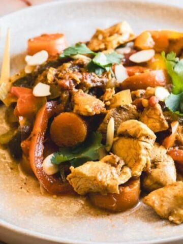 A plate of cooked chicken pieces with sliced carrots, bell peppers, and garnished with cilantro and almond slices. A fork rests on the side of the plate and an orange napkin is partially visible.
