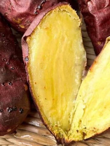 A woven basket contains several roasted sweet potatoes, with one sweet potato cut open to show its yellow, soft interior. The outer skin is reddish-brown and slightly wrinkled.