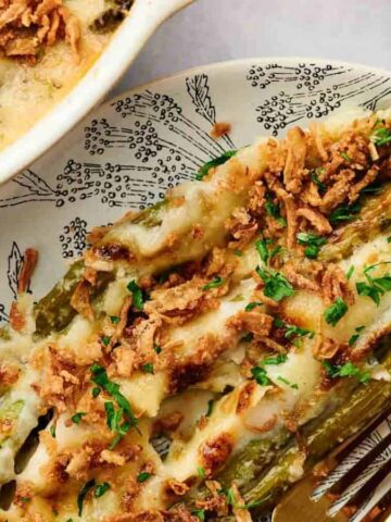 A plate with a serving of baked asparagus topped with melted cheese, crispy fried onions, and chopped herbs. A fork is beside the food, and a baking dish with more casserole is partially visible.