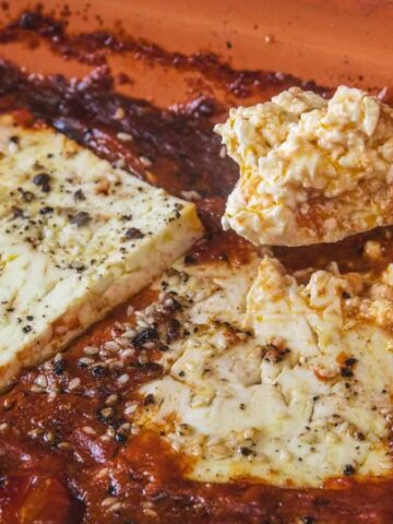 A close-up of baked feta cheese with cherry tomatoes in a rectangular baking dish. A spoon holds a serving of the crumbled, cooked feta above the dish. The cheese is seasoned with pepper and herbs.