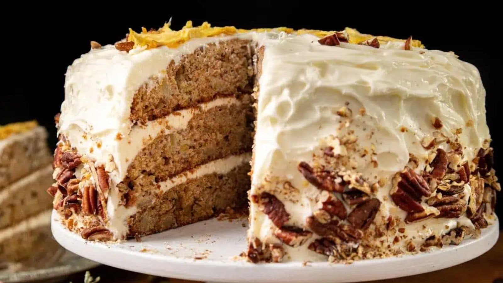 A slice of Hummingbird Cake with cream cheese frosting and pineapple chunks on top, placed on a decorative green and white plate. The Southern cake appears moist with visible nuts, and a fork is resting at the base.