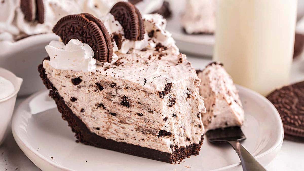 Slice of Oreo cream pie on a white plate, topped with whipped cream and Oreo cookies, with a fork resting beside it.