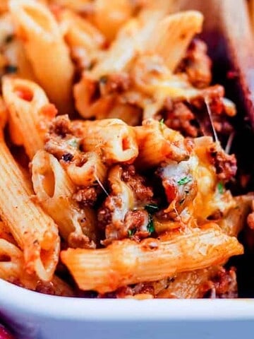 A close-up of baked penne pasta with ground meat and melted cheese in a white casserole dish, with a wooden spoon serving a portion.