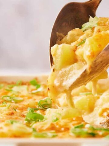 A metal serving spoon lifts a portion of cheesy potato casserole from a white baking dish. The casserole is topped with melted cheese and sliced green onions.