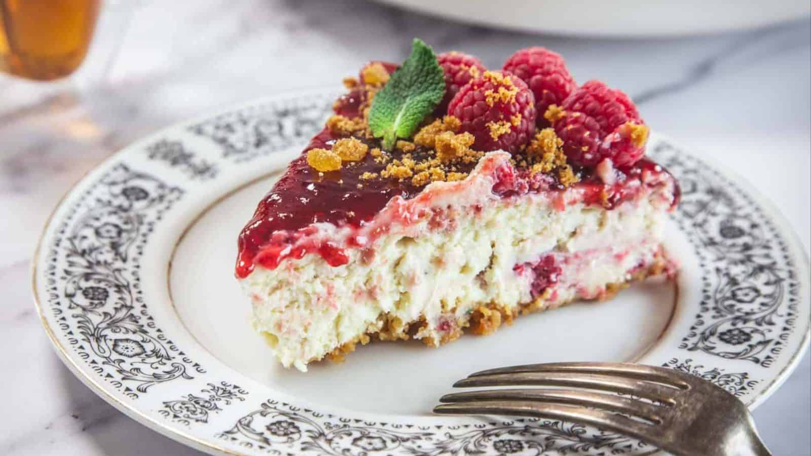 A slice of raspberry cheesecake topped with raspberries, mint leaves, and crumbled topping is placed on a decorative plate with a fork beside it.