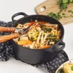 A black pot filled with Thai Red Curry noodle soup, tofu, bok choy, and vegetables sits on a patterned cloth with wooden-handled utensils inside. A cutting board with chopped herbs is nearby, completing the recipe scene.