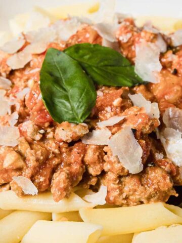 A white bowl filled with penne pasta topped with meat sauce, grated parmesan cheese, and fresh basil leaves, with a metal spoon resting on the side of the bowl.