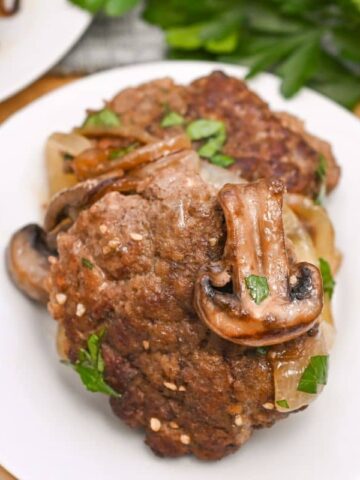 A white plate with a serving of cooked Salisbury steak topped with sautéed mushrooms and onions, garnished with chopped parsley. Fresh parsley is visible in the background on a wooden surface.