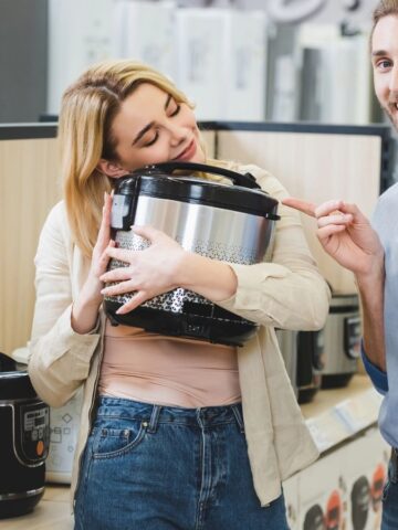 A woman holds and smiles at a pressure cooker in an appliance store, while a man beside her points at the appliance. Several other pressure cookers are displayed on the shelf.