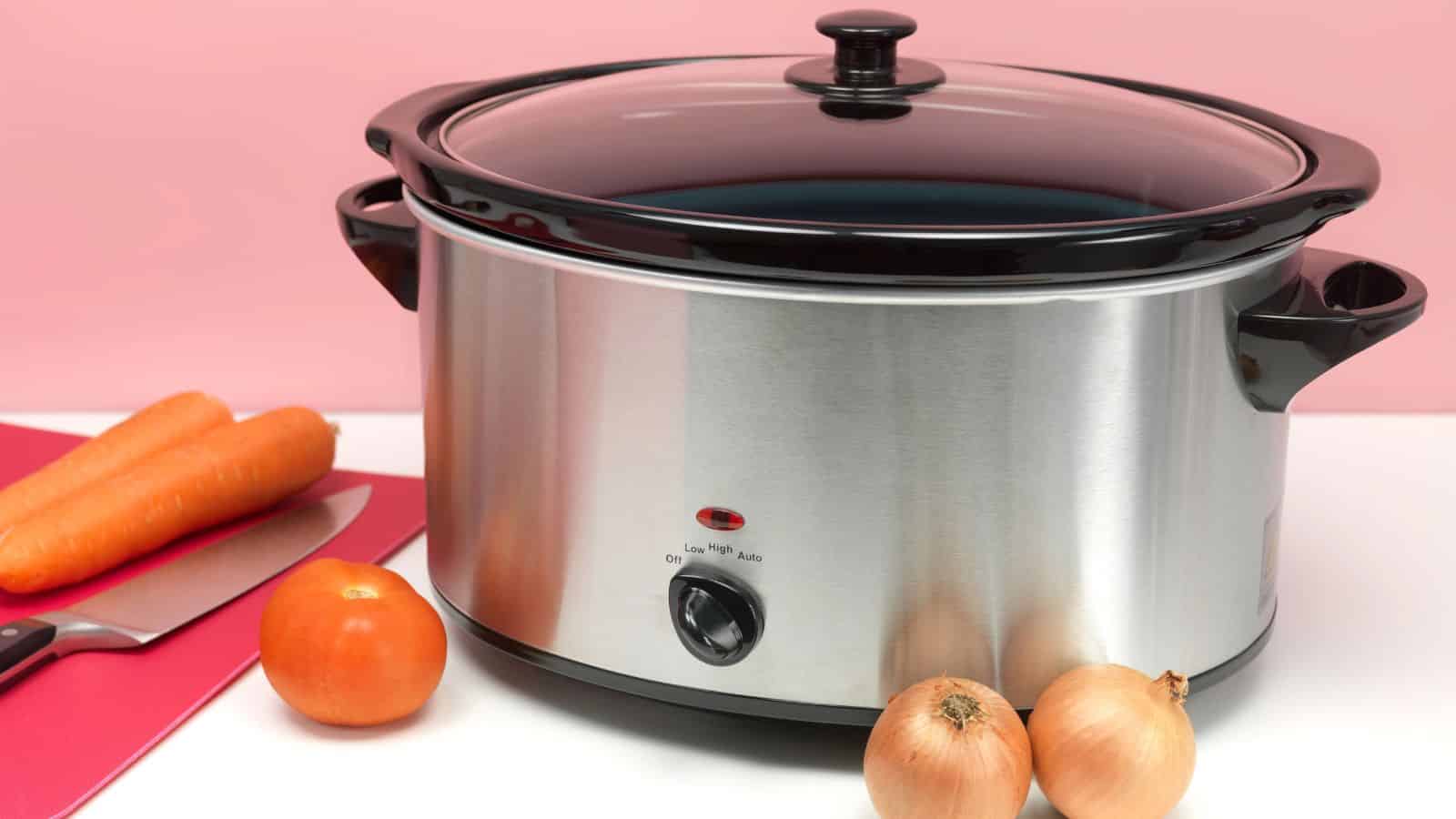 A stainless steel slow cooker with a black lid stands on a white counter. In front of it are two onions and a tomato. On the left, two carrots and a knife are on a red cutting board. The background is pink.