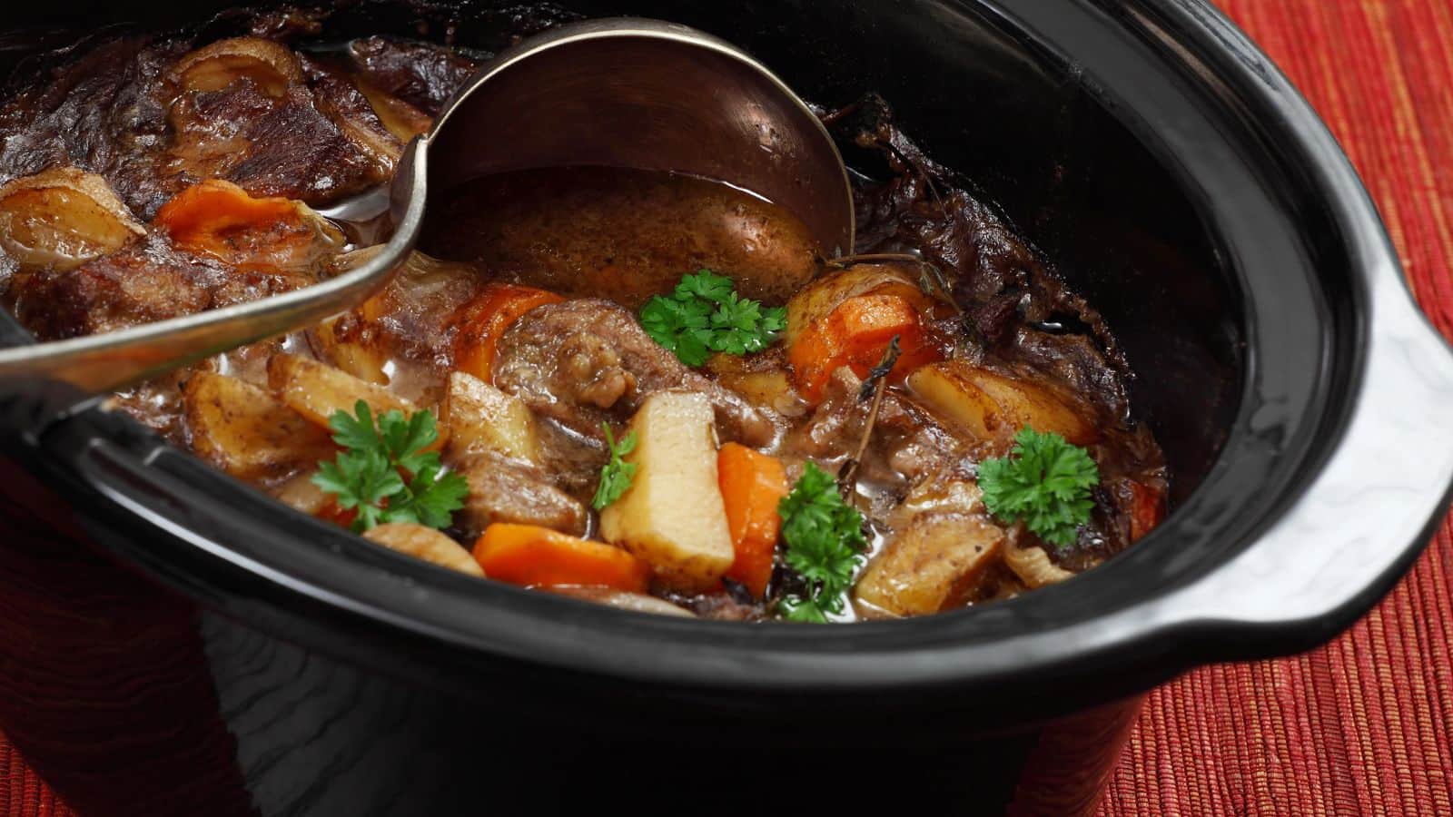 A slow cooker filled with beef stew, including chunks of beef, carrots, potatoes, and parsnips, garnished with fresh parsley. A metal ladle rests inside the slow cooker.