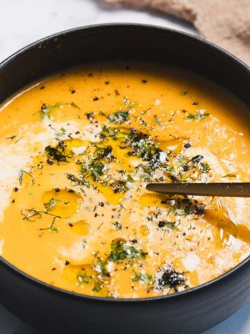 A black bowl filled with creamy orange soup, garnished with herbs, pepper, and a drizzle of oil. A gold spoon rests inside the bowl. A textured brown cloth and a cup are in the background.
