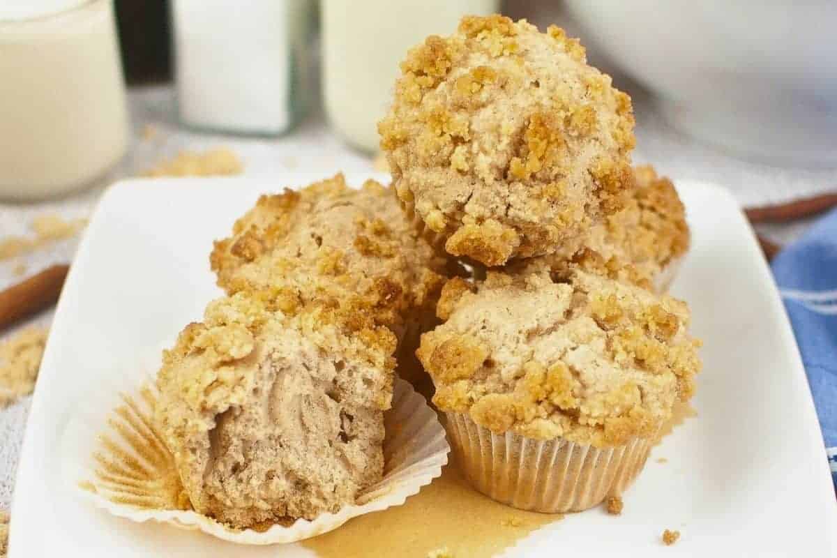 A plate of five cinnamon muffins with crumbly tops; one muffin partially unwrapped and cut in half.