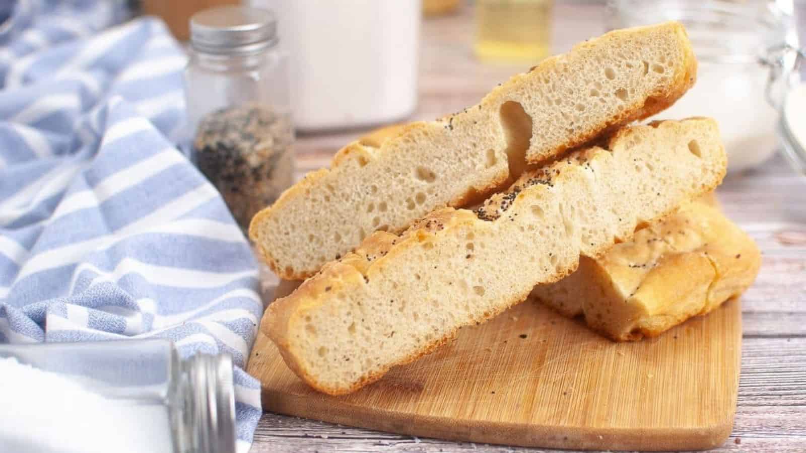Two slices of sourdough focaccia rest on a wooden cutting board, accompanied by a blue striped cloth and an array of seasoning containers in the background.
