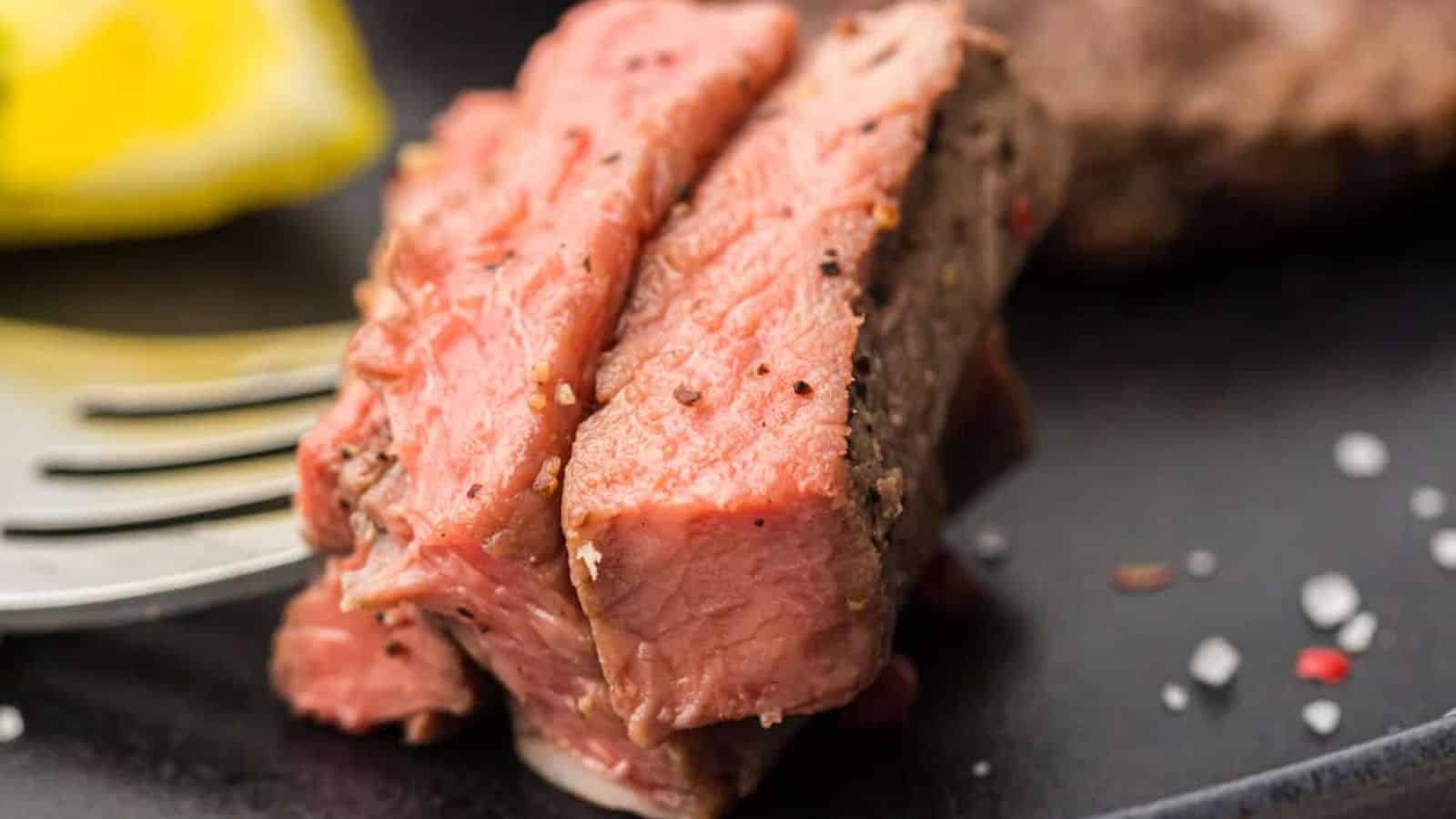 Close-up of a fork holding a piece of medium-rare cooked beef steak, with visible seasonings and a slice of lemon in the background.