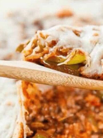 A wooden spoon holds a serving of casserole with melted cheese, green bell peppers, rice, and tomato sauce over a baking dish filled with the same mixture.