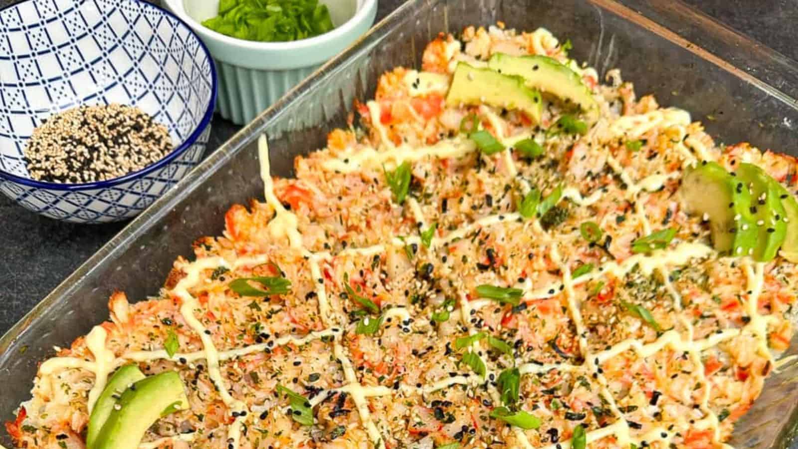 A baking dish filled with sushi bake, featuring layers of rice, seafood topping, mayonnaise drizzle, avocado slices, sesame seeds, and herbs.