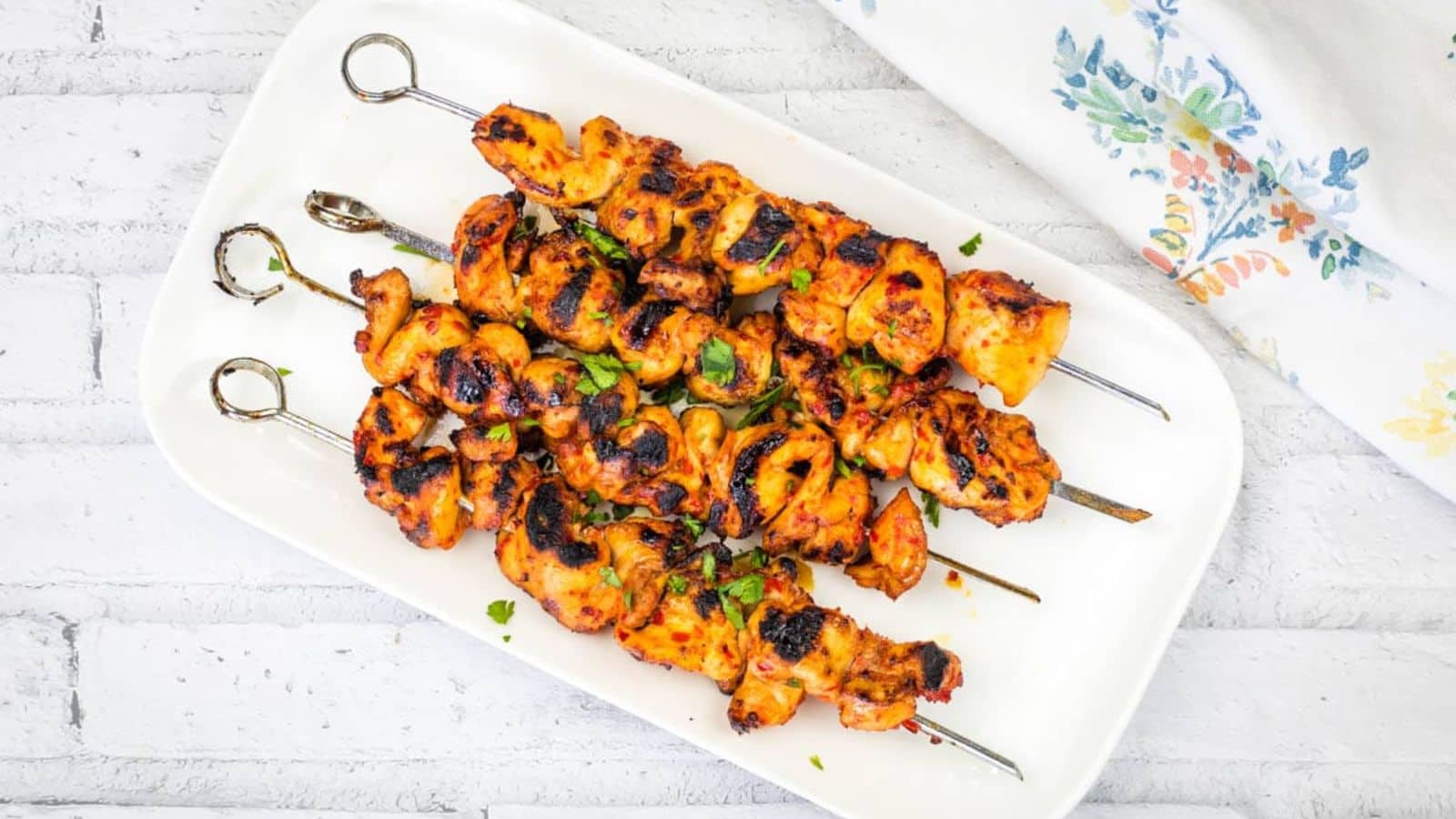 A rectangular white plate with four Sweet & Spicy Grilled Harissa-Honey Chicken Skewers garnished with chopped herbs.