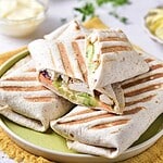 Grilled wraps filled with sliced chicken shawarma, lettuce, cucumber, and other vegetables are stacked on a green plate. A bowl of sauce and some herbs are visible in the background.