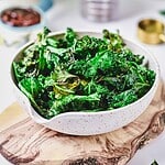 A white speckled bowl filled with crispy kale chips sits on a wooden serving board, featuring a tasty air fryer kale chips recipe, with blurred kitchen items visible in the background.